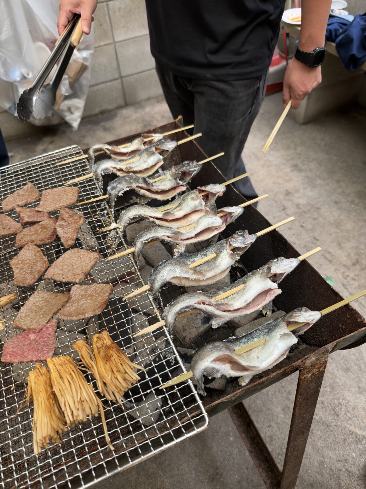 炭火の網で串に刺したマスと肉を焼くBBQの様子
